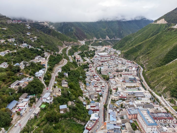 An aerial view of a Deqin County, surrounded by lush greenery and surrounded by mountains. The town is densely populated with houses and buildings, and the roads crisscross the valley. The town is surrounded by trees and hills, creating a serene and picturesque scene.