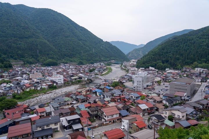 A serene mountain town with a river, houses, and buildings, surrounded by lush green mountains and trees.