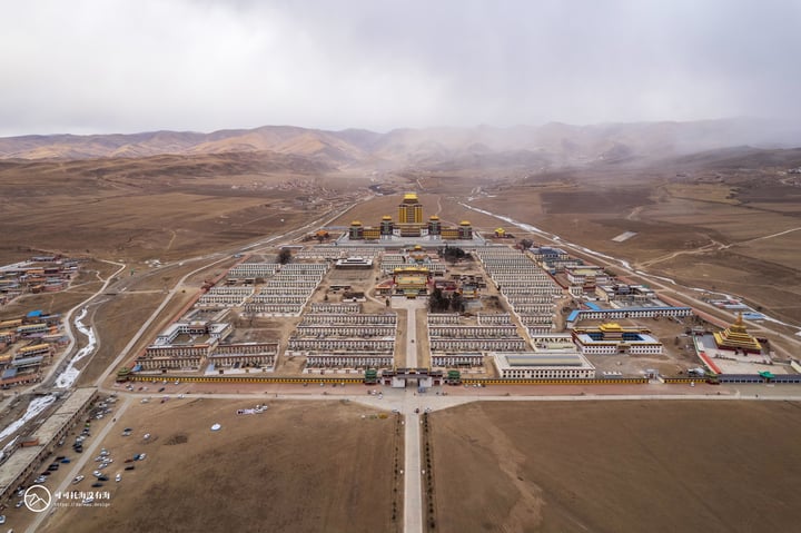 An aerial view of a large, beige and yellow complex with a central golden dome, surrounded by a barren landscape of brown hills and mountains, under a cloudy sky.