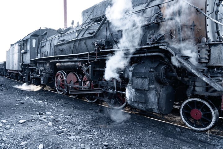 An old black steam locomotive with red wheels is traveling on a railway track, emitting smoke from its chimney.