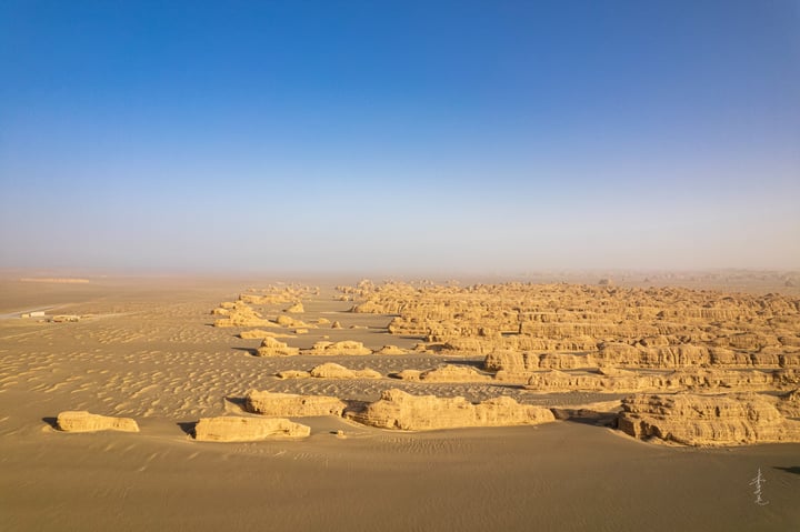 Aerial view of a desert landscape with sandy formations, a clear blue sky, and a few distant structures.