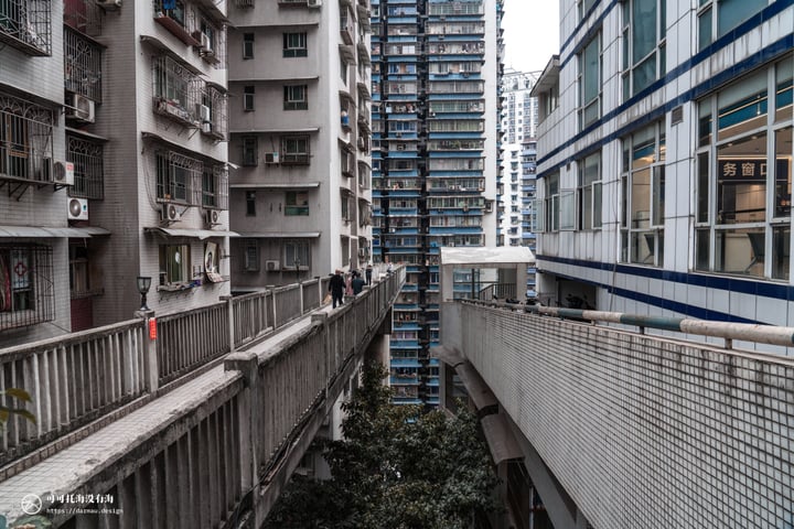 A cityscape with tall buildings, balconies, and windows, including a balcony with a red sign. Trees and a fence are visible in the foreground.