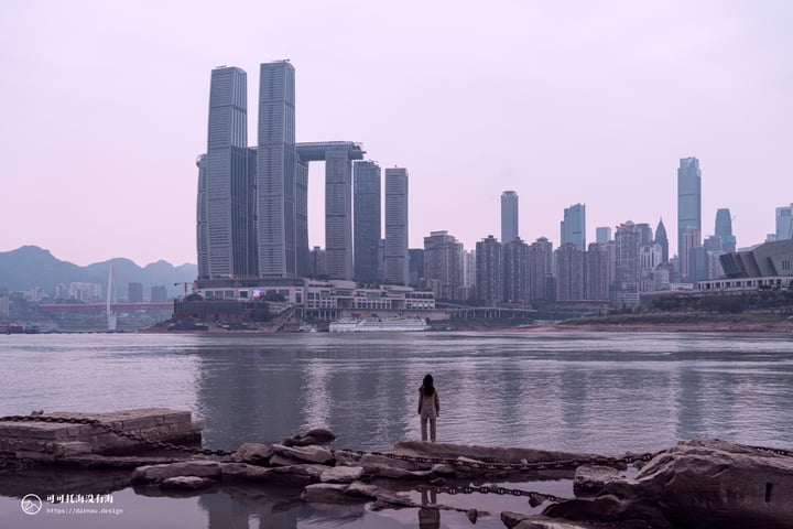 A person stands on rocks near a calm body of water, with a city skyline in the background and a red bridge over the water.