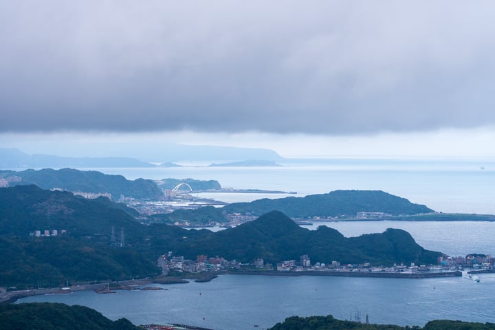 A coastal city with a harbor, mountains, and a cloudy sky, with boats on the water and a distant island.