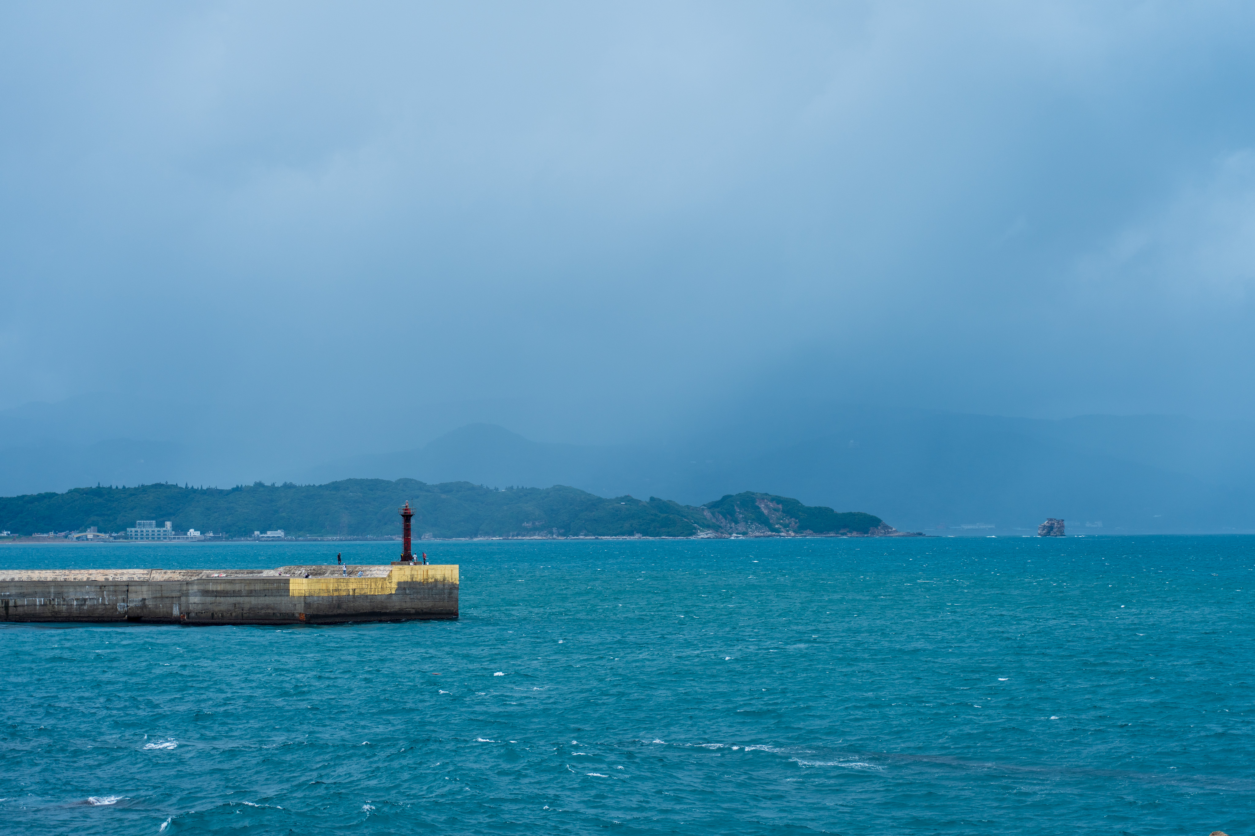 A lighthouse stands on a concrete pier in the ocean, with a mountain range in the distance and a stormy sky overhead.