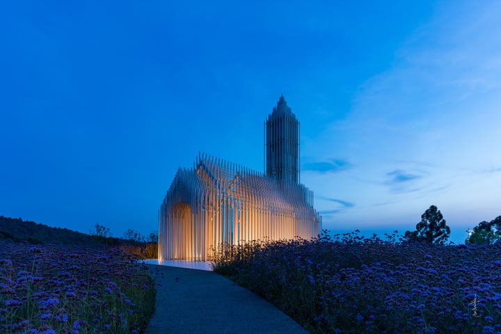 A modern church with a tall tower and glass facade stands in a field of purple flowers, illuminated by a pathway leading up to it. The sky is dark blue with a few clouds, and the scene is serene and peaceful.