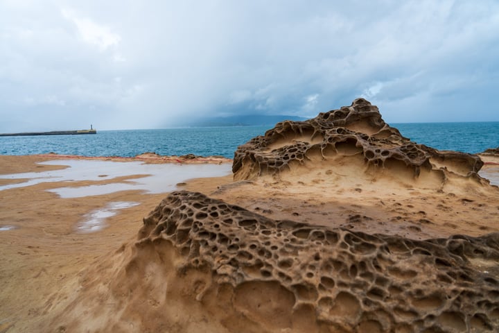 A rocky beach with a large, textured rock formation in the foreground, a calm ocean with a lighthouse in the distance, and a cloudy sky above.