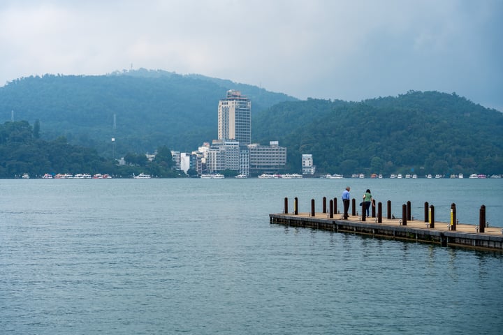 A wooden pier extends into a calm body of water, with a few boats and a few people standing on it. In the distance, a city with a tall building and a mountain range are visible.