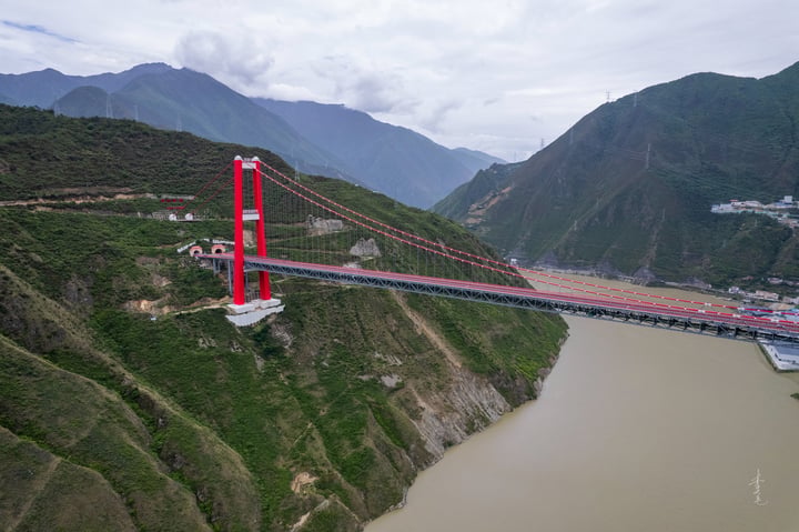 A red and white suspension bridge spans a river in a valley, with mountains in the background. The bridge has a white base and red pillars, and is supported by red cables. The river is brown and has a boat on it. The sky is cloudy.