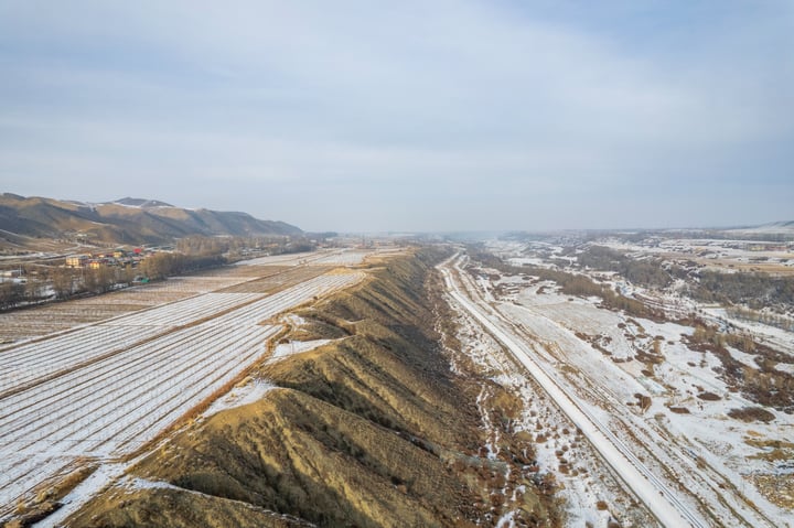 A snowy landscape with mountains and hills in the background, a road running through the middle, and a few buildings in the distance.