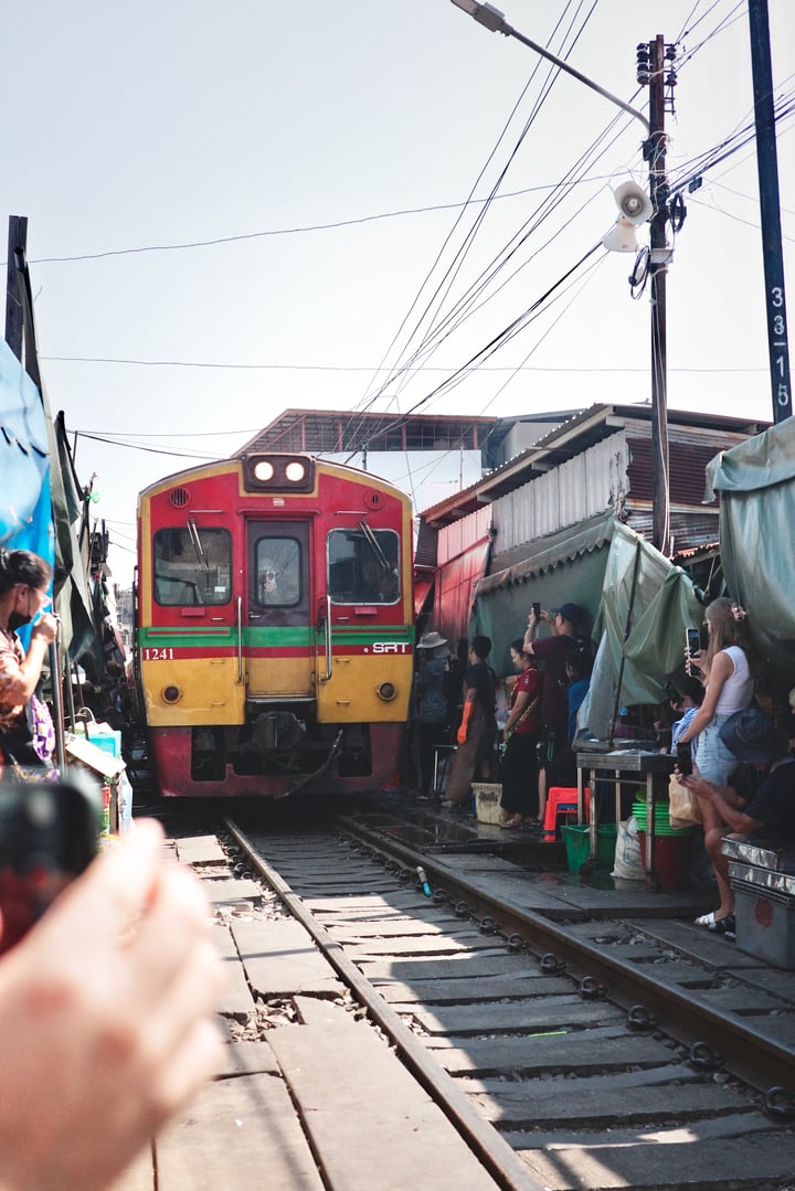 A red and yellow train is passing through a bustling train station, surrounded by people waiting to board or disembarking.