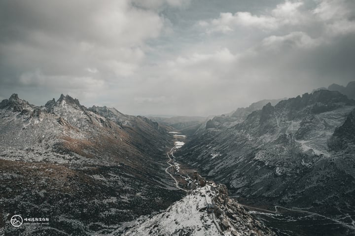 A snowy mountain range with a winding river in the valley, surrounded by a cloudy sky.