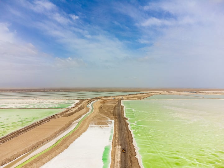 An aerial view of the Lake of the Dead in Bolivia, showcasing its vibrant green and turquoise waters, surrounded by a sandy beach and a dirt road. The sky is a clear blue with a few clouds, creating a serene atmosphere.