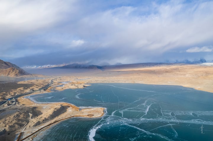 A serene landscape features a deep blue lake reflecting the sky, with snow-capped mountains in the distance. The sky is filled with white clouds, and the landscape is dominated by brown and beige tones.