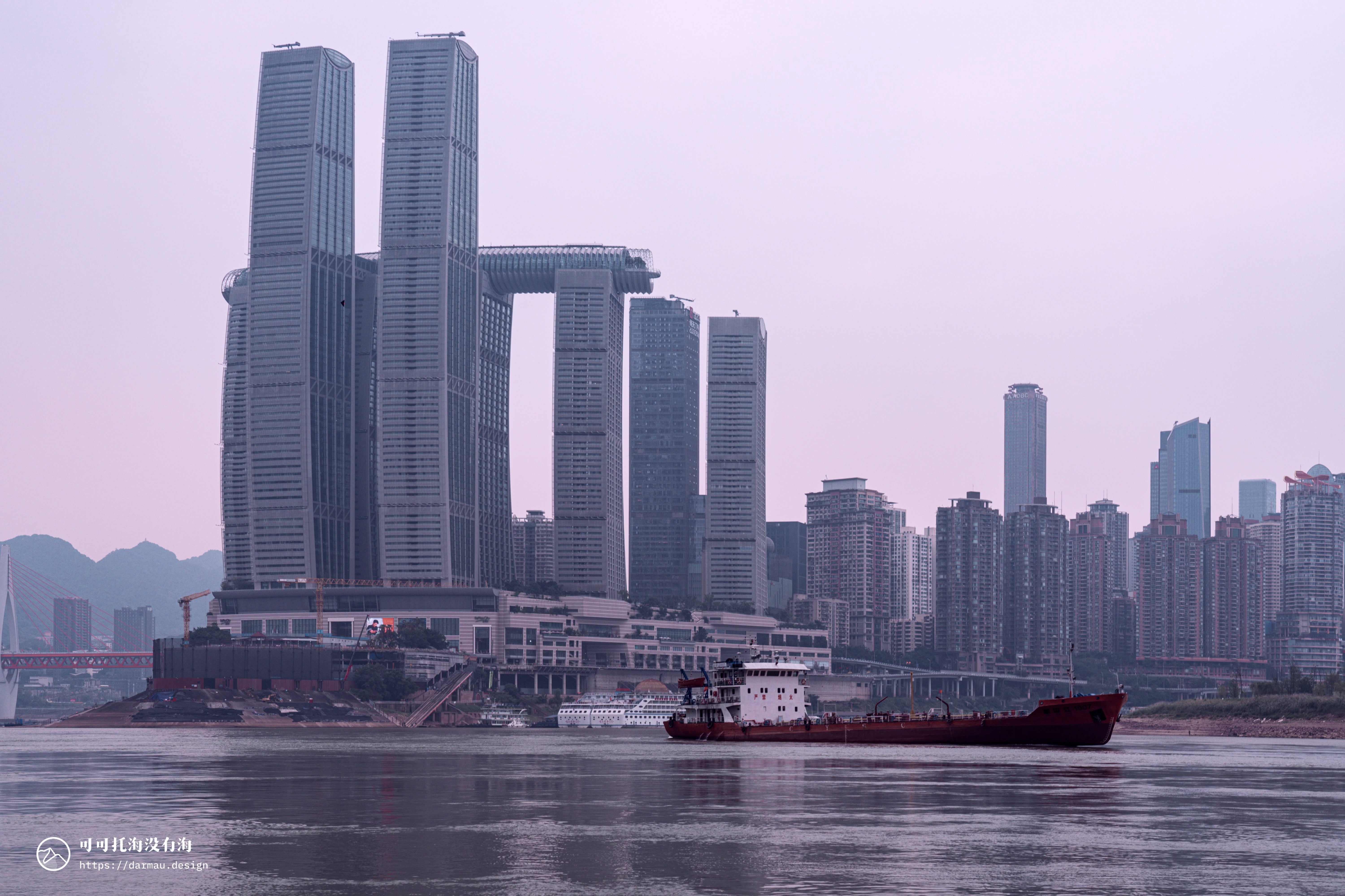 A cityscape with tall buildings, a river, mountains, and a red crane.