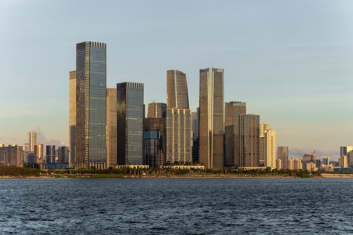 A cityscape with tall buildings, including skyscrapers, is captured in a serene, clear sky. The water in the foreground is calm, and the buildings are scattered across the water, creating a dynamic urban scene.