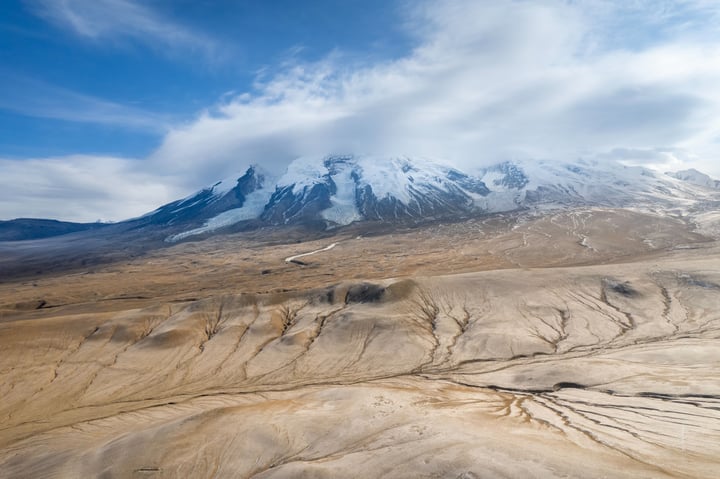 A snow-capped mountain range dominates the landscape, with a vast, sandy desert stretching out in the foreground. The sky is a mix of blue and white, with clouds scattered across it. The image is taken from an aerial perspective, emphasizing the grandeur of the mountain range and the vastness of the desert.