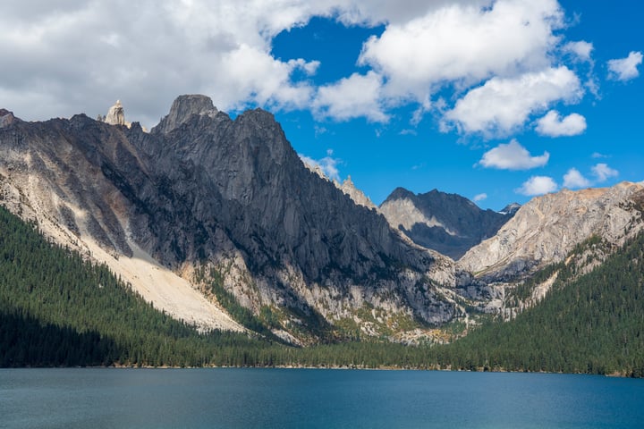 A serene mountain lake surrounded by towering gray and brown mountains, with a clear blue sky and fluffy white clouds.
