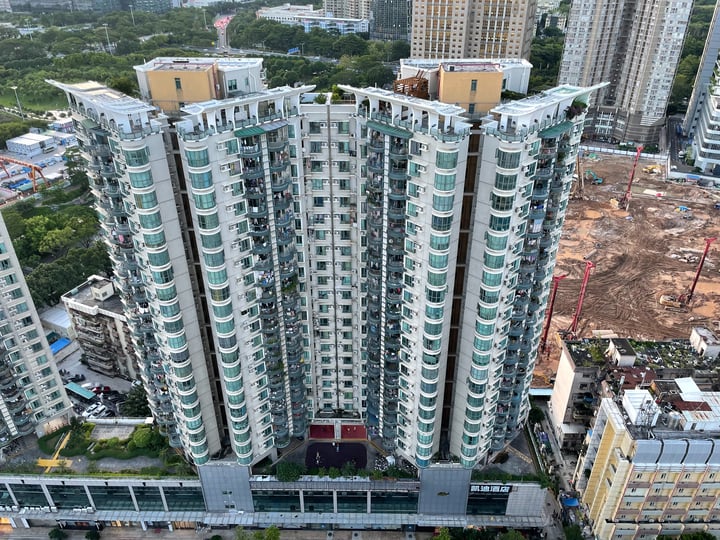 An aerial view of a tall, white apartment building with balconies and windows, surrounded by other buildings and construction sites, showcasing urban development and growth.