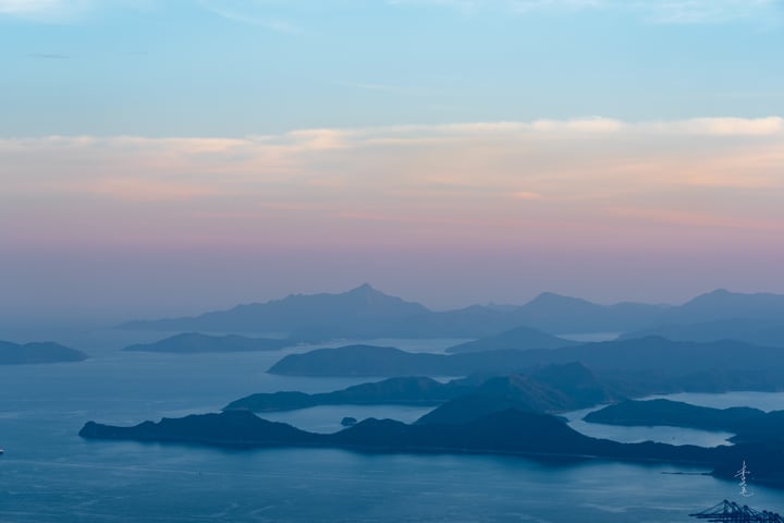 A breathtaking aerial view of a mountainous landscape with a large body of water in the foreground, surrounded by mountains and islands. The sky is a mix of blue and pink hues, with a few clouds scattered across. A small airplane is visible in the distance, adding a sense of scale and movement to the scene.