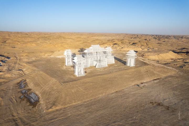 An aerial view of a white, pagoda-like structure with multiple levels and a central roof, surrounded by a barren desert landscape.