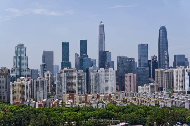 A cityscape with tall buildings, including a prominent skyscraper, is captured from a high vantage point. The sky is clear blue, and the buildings are predominantly gray and white. Trees are scattered throughout the foreground, adding a touch of nature to the urban landscape.