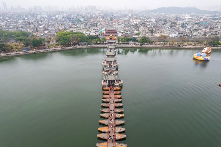 这是一张航拍图，显示了一座位于水面上的多层塔楼，通过一条用船连接的木桥与岸边相连。背景是密集的城市建筑和城市天际线，远处还有一个巨大的黄色充气鸭子漂浮在水中。
