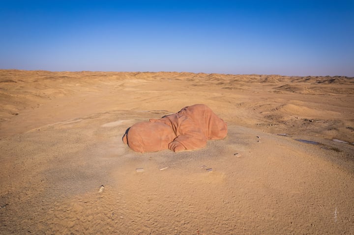 A large red rock is in the center of a sandy desert landscape, surrounded by sand dunes and a clear blue sky.