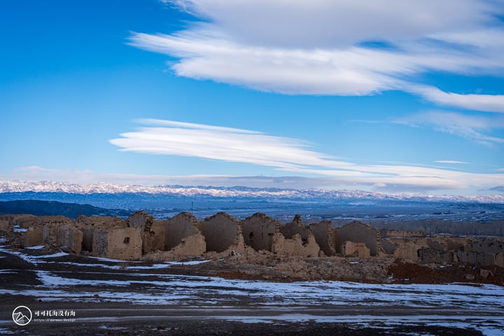 An old, weathered stone ruins stand in a barren landscape, surrounded by snow-covered mountains and a blue sky with white clouds.