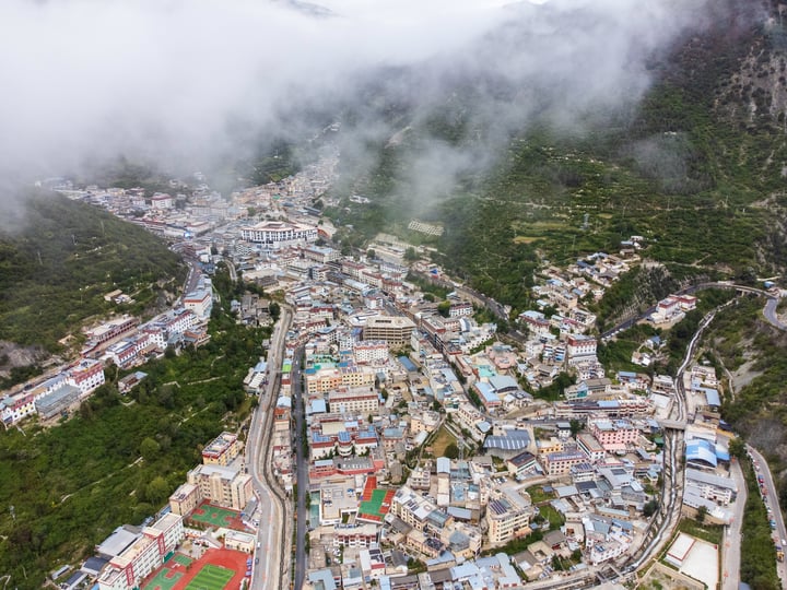 An aerial view of a Deqin County, surrounded by lush greenery and surrounded by mountains. The town is densely populated with houses and buildings, and the roads crisscross the valley. The town is surrounded by trees and hills, creating a serene and picturesque scene.