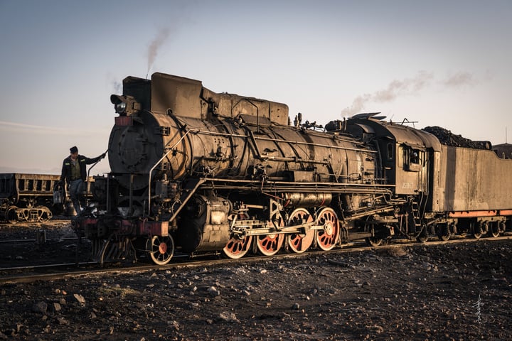 An old black steam locomotive with red wheels is traveling on a railway track, emitting smoke from its chimney.