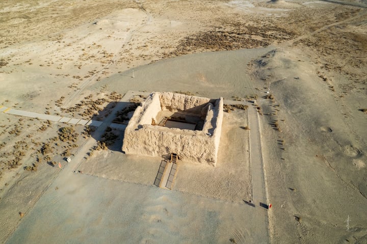 An aerial view of a large, rectangular stone structure in a desert-like environment, surrounded by sandy terrain and a few scattered rocks.