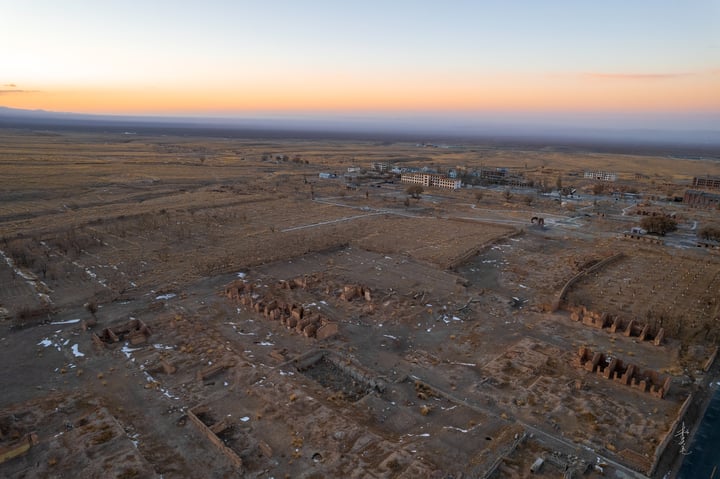 An aerial view of a desert landscape with a mix of buildings and structures, surrounded by a vast expanse of dry grass and a few scattered trees. The sky is a mix of orange and blue hues, indicating either sunrise or sunset.