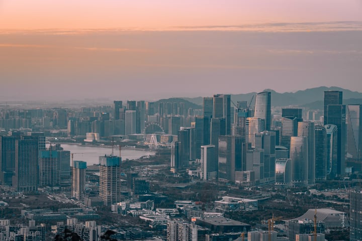 A cityscape at sunset, with tall buildings in shades of blue and gray, reflecting the orange and pink hues of the setting sun. A river runs through the city, and mountains are visible in the distance.