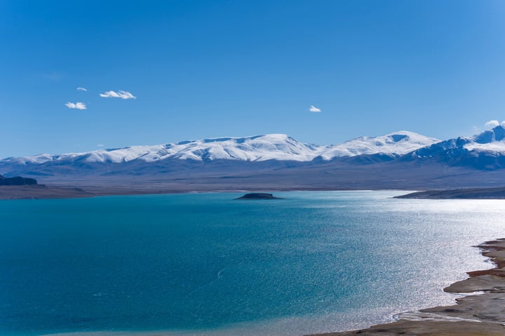 A breathtaking aerial view of a serene lake surrounded by snow-capped mountains, with a clear blue sky above.
