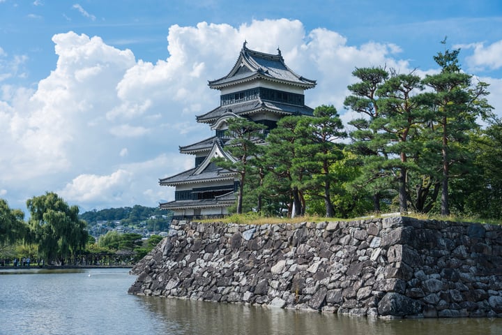 A serene scene features a traditional Japanese castle on a stone wall, surrounded by trees and a calm body of water. The sky is a clear blue with white clouds, and the castle's architecture is visible through the stone wall.