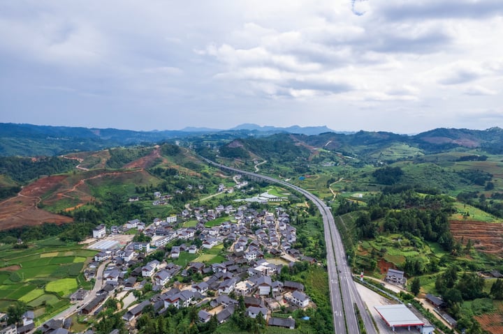 An aerial view of a small town nestled in a valley, surrounded by rolling hills and lush greenery. The town is densely populated with houses and buildings, and a winding road runs through it. The sky is filled with clouds, casting a soft light over the scene.