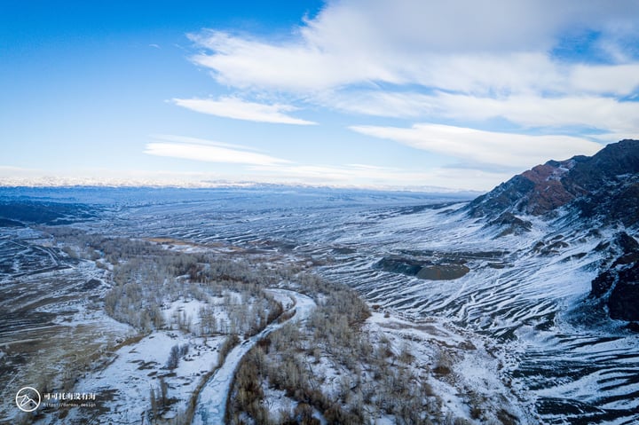 A snowy mountain landscape with a winding road, snow-covered trees, and a blue sky with clouds.
