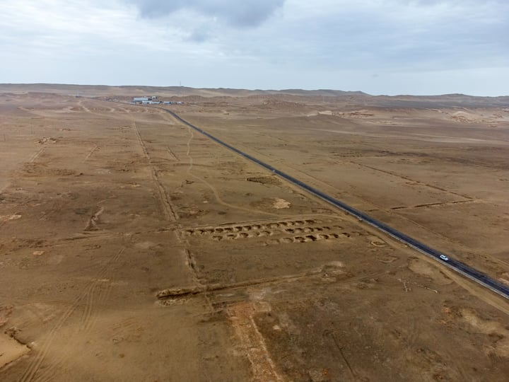 An aerial view of a barren desert landscape with a long, straight road stretching into the distance. The road is bordered by sandy dunes and is surrounded by a few buildings and a few cars. The sky is cloudy, casting a soft light over the scene.