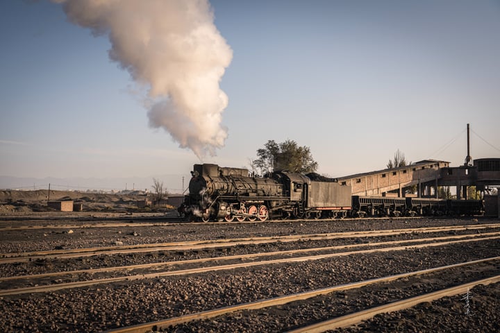 An old, black steam locomotive is on a set of train tracks, emitting white smoke, with a small building and trees in the background.