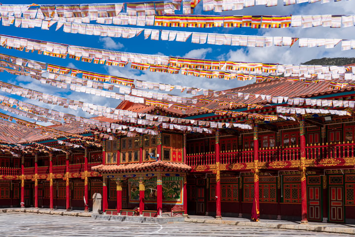 A traditional Chinese temple with red and gold walls, white and gold columns, and a red roof is surrounded by a blue sky with white clouds. The temple is in a courtyard with a wooden floor and a stone wall. The image is taken from a low angle, emphasizing the temple's grandeur.
