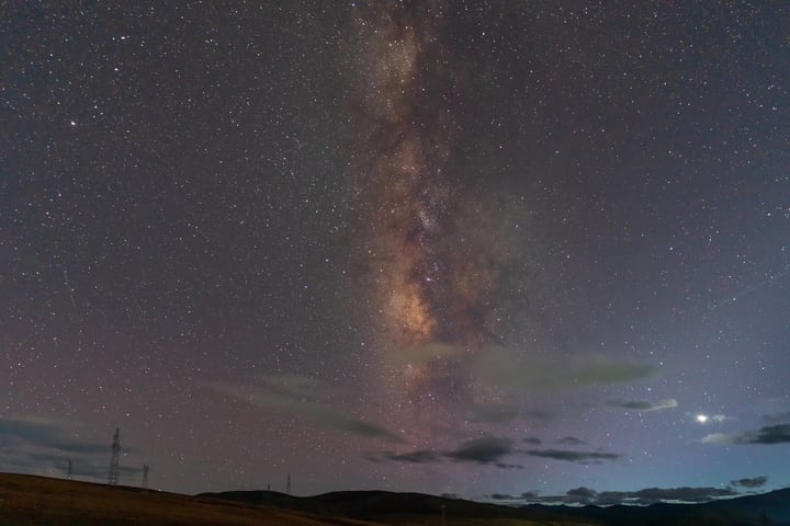 A serene night scene features a vast, star-filled sky with a few scattered clouds, a bright, glowing Milky Way, and a dark, grassy field with power lines.