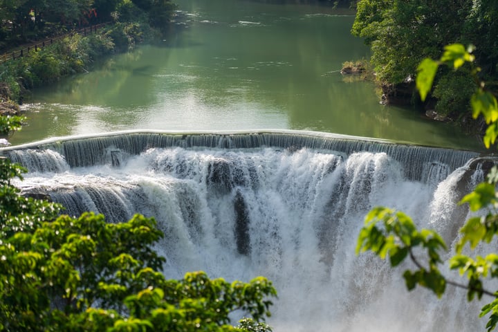 A large waterfall cascades over a body of water, surrounded by lush greenery and a clear blue sky.