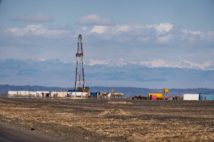 A tall, red and white oil rig stands in a barren, rocky landscape, with a mountain range in the distance.