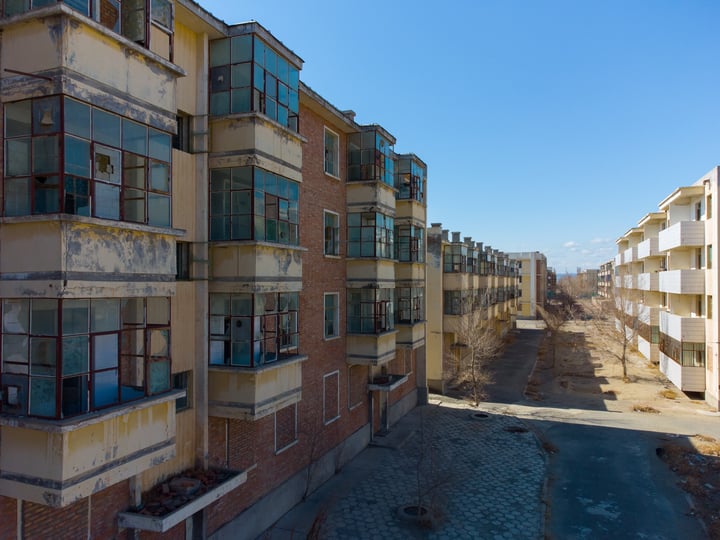 A row of old, multi-story buildings with a mix of brick and concrete, some with balconies, are lined up on a street. The buildings have a mix of colors and are surrounded by trees. The sky is clear blue.