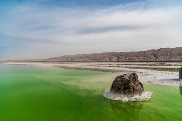 A serene desert lake with a bright green color contrasts against a sandy beach and rocky shore, surrounded by a mountain range under a clear blue sky.