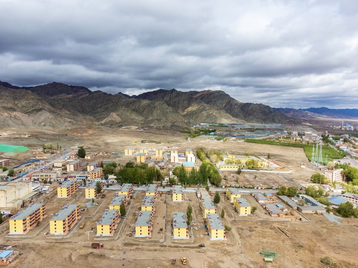 An aerial view of a construction site with numerous buildings, surrounded by mountains and a bridge, with a large body of water in the background.