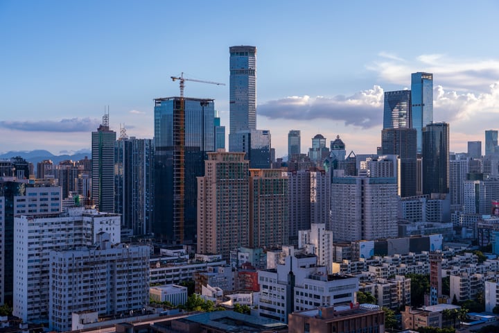 A city skyline at dusk, with a prominent skyscraper, a curved building, and a mountain range in the background.