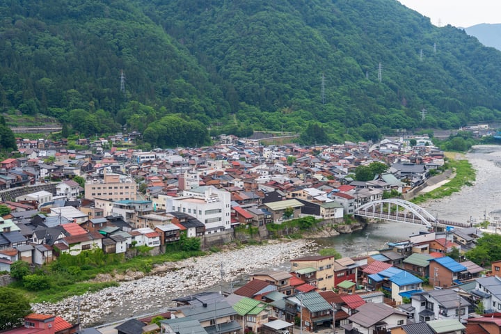 An aerial view of a town nestled on a hillside, surrounded by lush green mountains and a tranquil river. The town is densely packed with colorful houses, and a white bridge spans the river. The sky is overcast, casting a soft light over the scene.