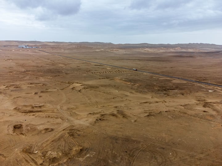 An aerial view of a barren desert landscape with a road running through it, surrounded by sandy dunes and mountains.